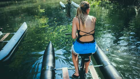 Woman wearing a black swimsuit and blue shorts sits on a water bike in the river