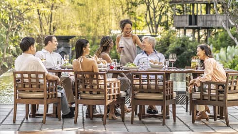 A long brown table with a family sat at it, in front of large trees.