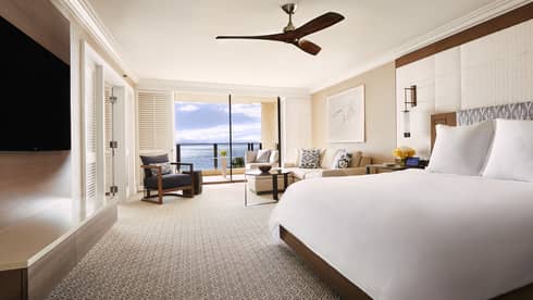 Bright suite with king-size bed, seating area and ocean-view balcony, featuring neutral tones and a ceiling fan at a Four Seasons luxury resort
