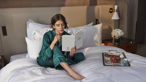 Person in green pajamas reading a book on a bed with a breakfast tray, including eggs and toast, in a cozy hotel setting