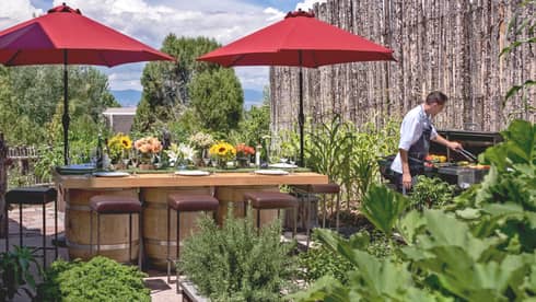 Chef grills vegetables, meats by long patio dining table, red umbrellas