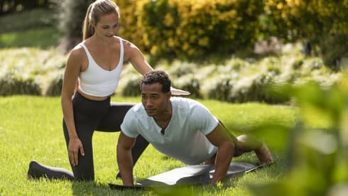 A yoga instructor helping a man get into a yoga position outside on very green grass.