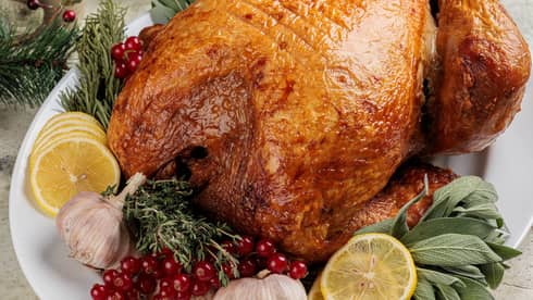 Whole roasted turkey on a white platter with lemon slices, green leaves, cranberries, garlic cloves and an assortment of holiday dishes in the background