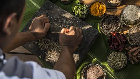 Photo of hand crushing spices on chopping spices, bowls of colourful spices and dried herbs around