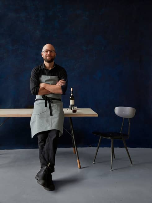 Portrait photo of Chef Greg Vernick leaning against table with bottle of wine