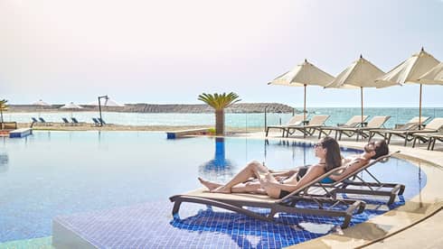 A couple lounges by the pool with view of ocean, beach umbrellas 