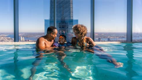 A father, mother and son in a pool surrounded by large windows looking out at a city skyline.