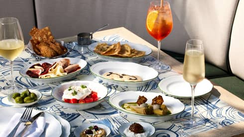 Outdoor table filled with a variety of food on white plates and three stemmed glasses filled with wine and cocktails