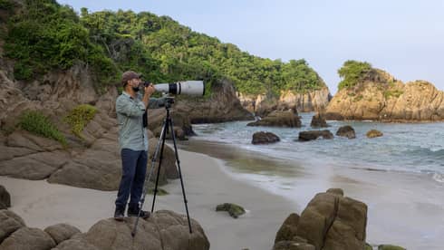 Photographer stands behind large camera lens on a rocky beach