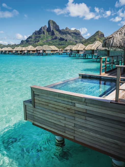 Overwater bungalows with thatched roofs on a turquoise lagoon, set against a mountain backdrop under a clear blue sky.