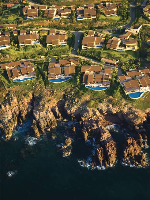 Aerial view of ocean, rugged cliffs and resort casita roofs and blue swimming pools, connecting roads