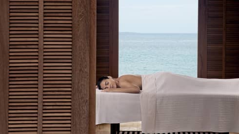 Guest enjoying a relaxing outdoor massage with a serene ocean view, framed by wooden shutters at a luxury beachfront resort
