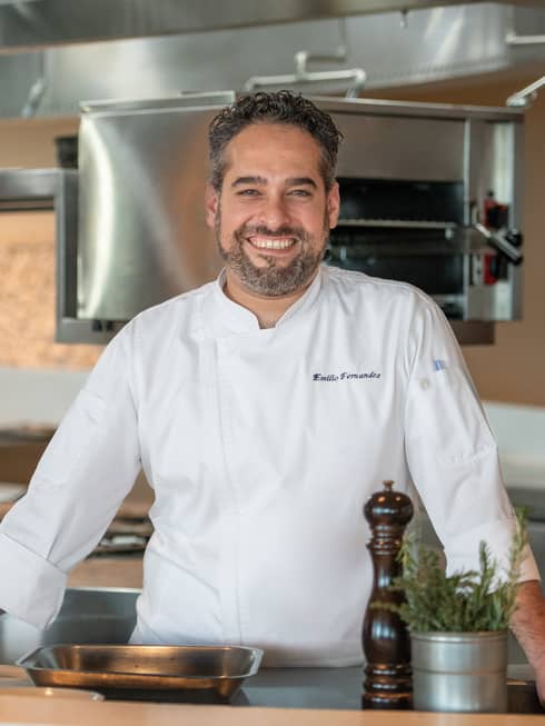 Executive Chef Emilio Fernandez in white chef's coat, standing in restaurant kitchen, smiling