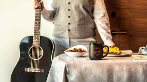 Person standing next to a black acoustic guitar, with a breakfast spread on a table featuring a "Gibson Garage" mug, fruit bowl, and scrambled eggs on toast.