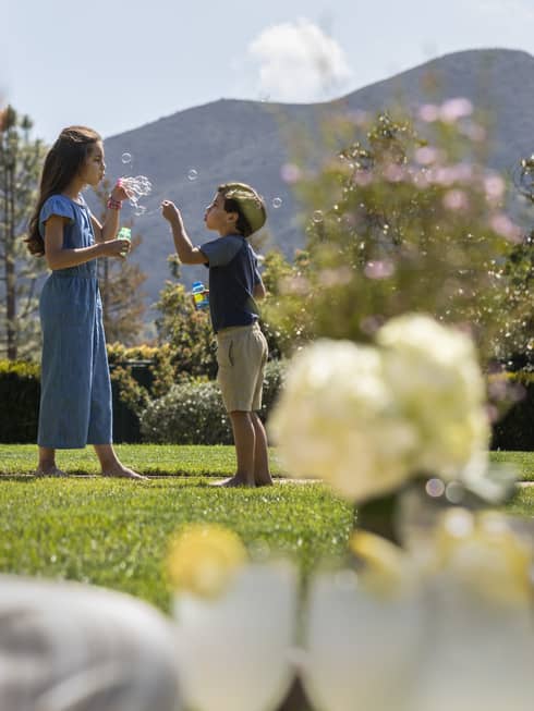 Two children blowing bubbles outside in the distance while a woman drinks a glass of wine in the foreground.
