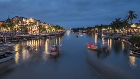 Evening view of a bustling riverside town with boats moving on the water, illuminated by streetlights and buildings along the waterfront, under a deep blue sky.