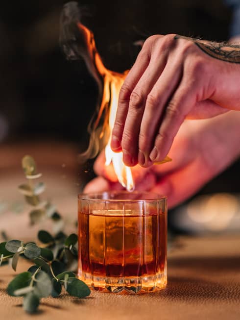 Bartender's tattooed hands light a flame above a cocktail served in a rocks glass next to a sprig of fresh eucalyptus