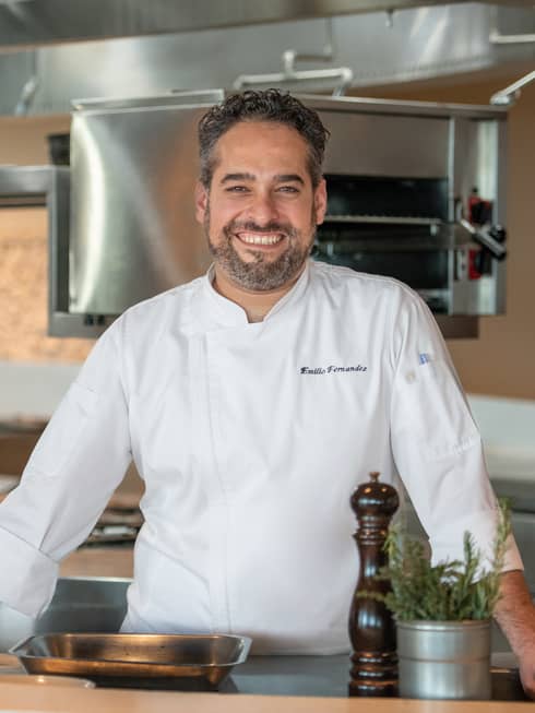 Executive Chef Emilio Fernandez in white chef's coat, standing in restaurant kitchen, smiling