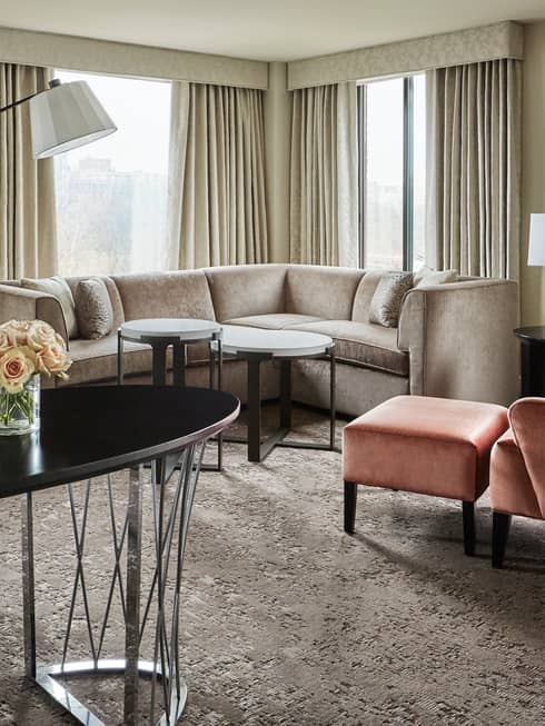 Capital Suite living room with long oval work desk in front of L-shaped sofa, pink armchair, corner windows