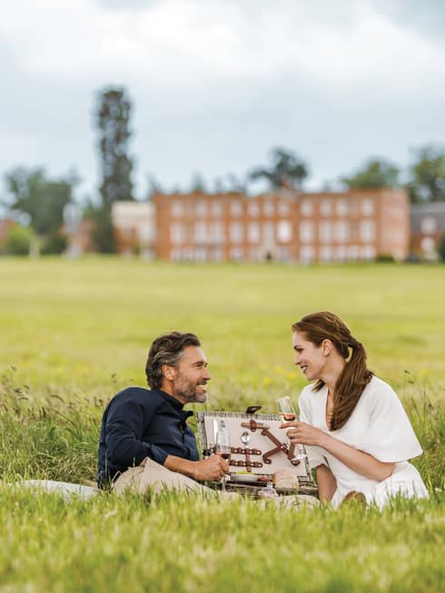 Couple with wine glasses lies in tall green grass with open picnic basket, hotel manor in background