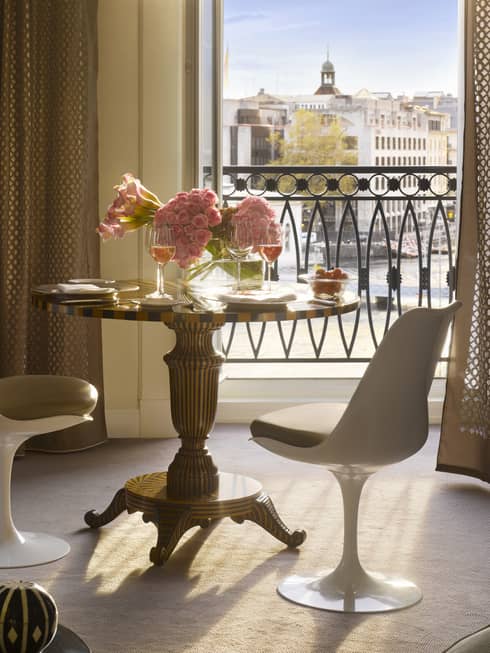 Round dining table and white bucket chair set with fresh pink flowers, wine glasses in front of sunny French balcony