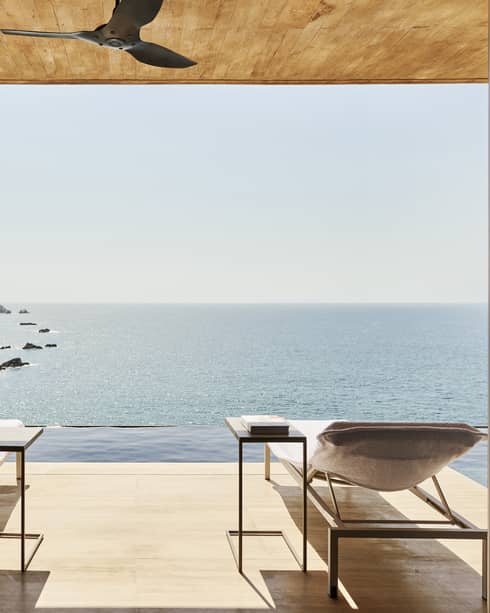 Open terrace with two lounge chairs and side tables, overlooking an infinity pool and a stunning ocean view with rocky islands. The space has a natural wood ceiling and serene ambience.