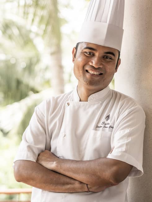Smiling person in white chef's jacket and hat stands with arms crossed on balcony