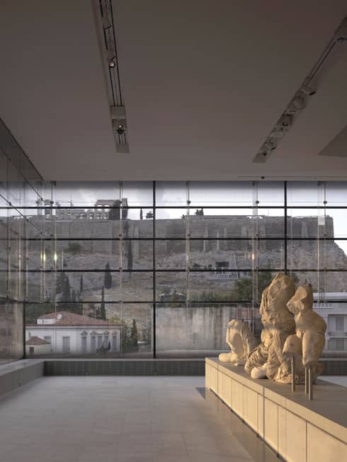 Greek sculptures on display in glass room overlooking ruins at Acropolis Museum