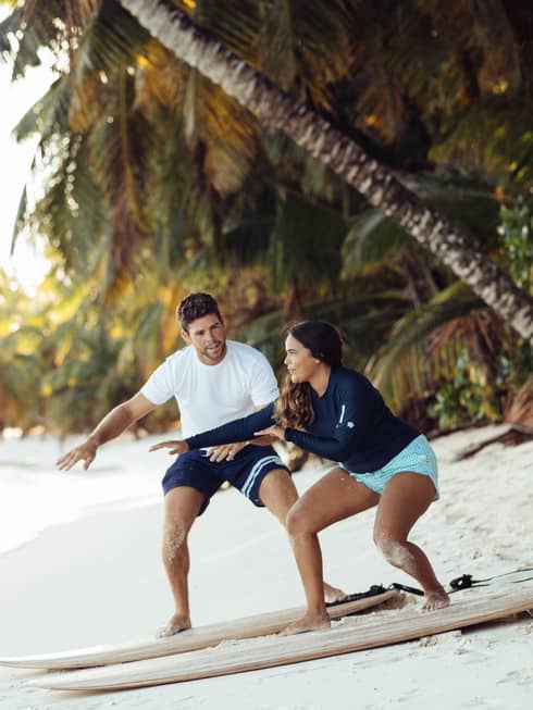 Man shows woman a surfing pose as they balance on surfboards on white sand beach under palm tree