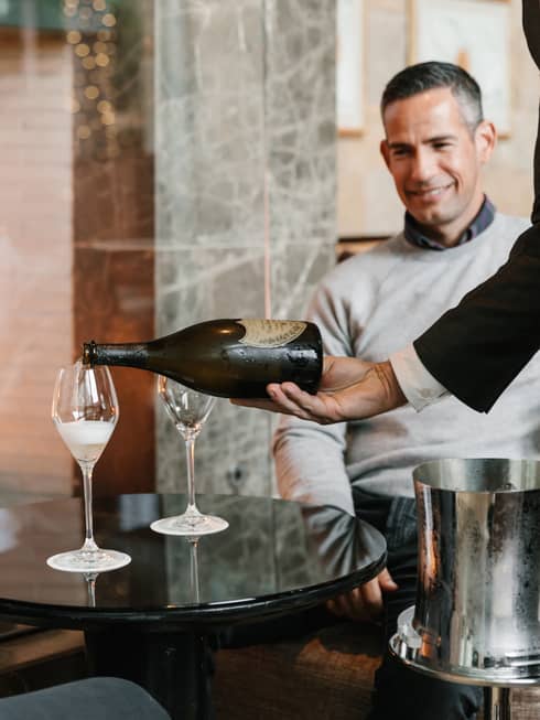 Two men being poured a glass of wine while sitting next to a fire.