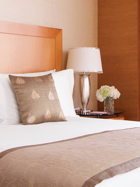 Embroidered accent pillows on hotel bed beside silver lamp, nightstand, flowers