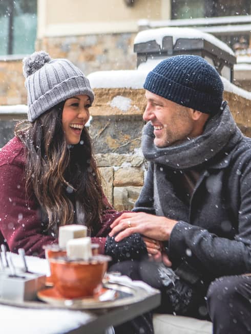 Smiling couple sitting by a firepit in a snowy outdoor setting, bundled in winter coats and hats. Snowflakes fall around them as they enjoy a cosy moment. 