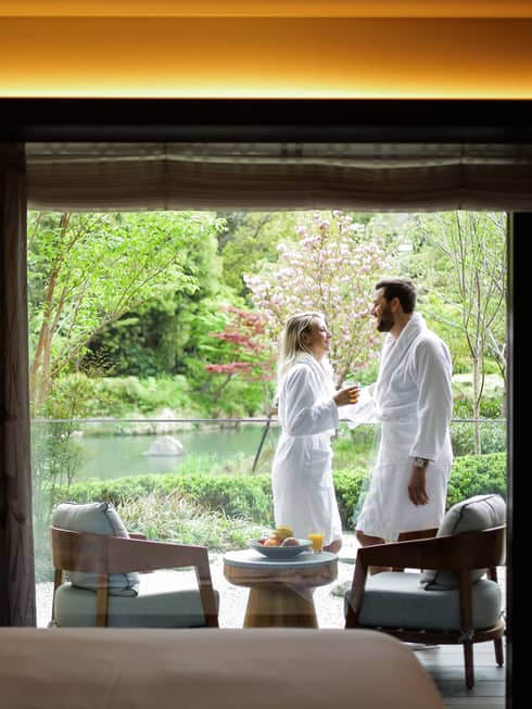 Smiling couple wearing white bathrobes on sunny balcony past hotel room bed