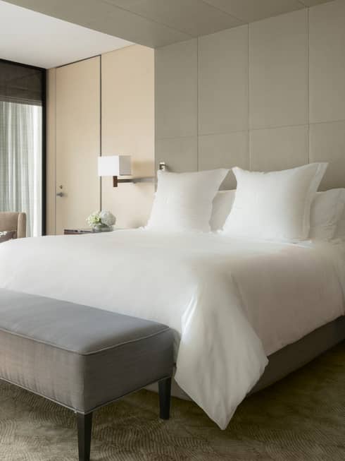 Sunny Deluxe One Bedroom Suite hotel bed with white linens, grey bench at foot