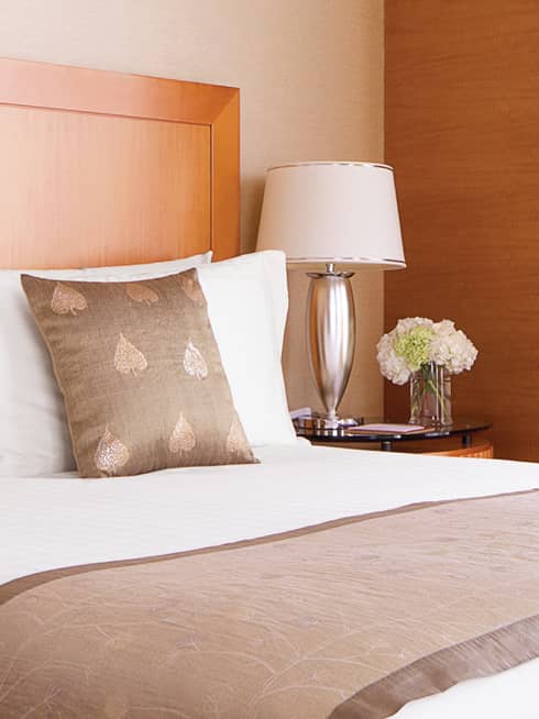 Embroidered accent pillows on hotel bed beside silver lamp, nightstand, flowers