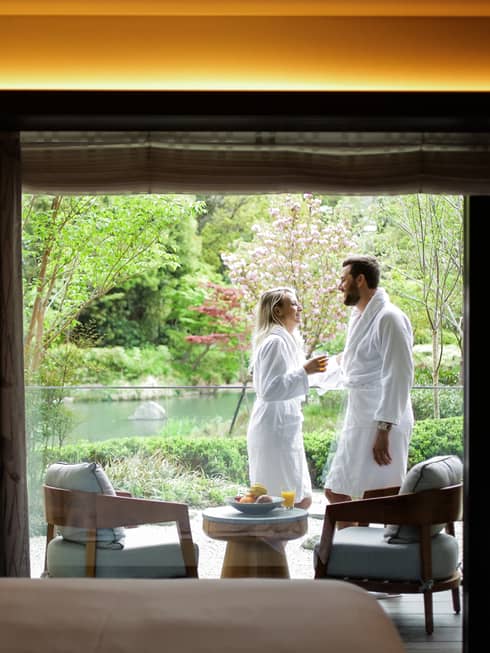 Smiling couple wearing white bathrobes on sunny balcony past hotel room bed