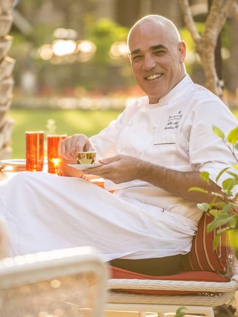Executive Chef Gilles Arzur smiling, in white uniform, sitting on patio chair, holding gold espresso cup