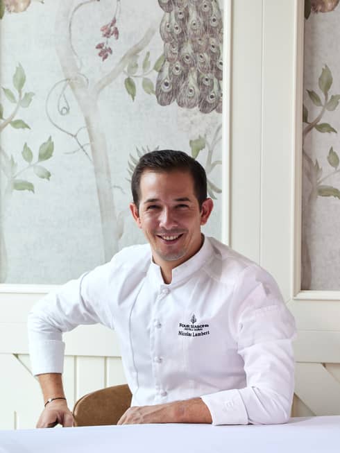 Chef wearing long-sleeve white chef's jacket sits at a table smiling, with floral wallpaper and white woodwork behind him