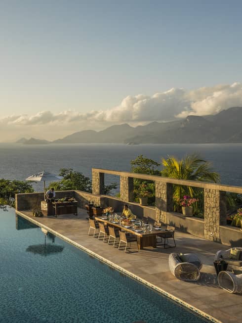 Six-Bedroom Residence Villa with long dining tables beside swimming pool reflecting a palm tree and the sunset. 