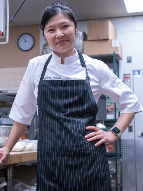 A female chef leaning on a counter in a kitchen with a blue apron on