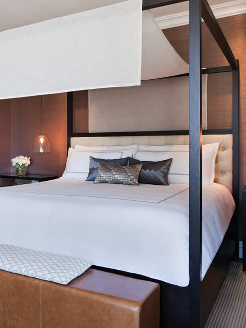Poster bed with white linen canopy, silk accent pillows, brown leather bench at foot