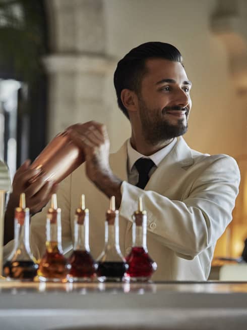 Smiling bartender wearing suit and tie shakes cocktail shaker, bottles with coloured liquid in foreground
