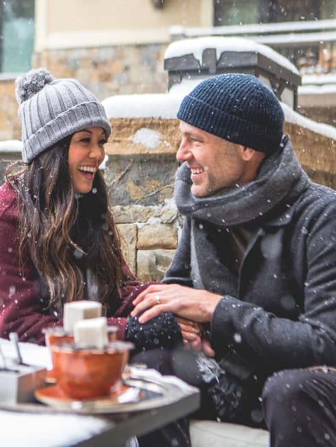 Smiling couple sitting by a firepit in a snowy outdoor setting, bundled in winter coats and hats. Snowflakes fall around them as they enjoy a cosy moment. 