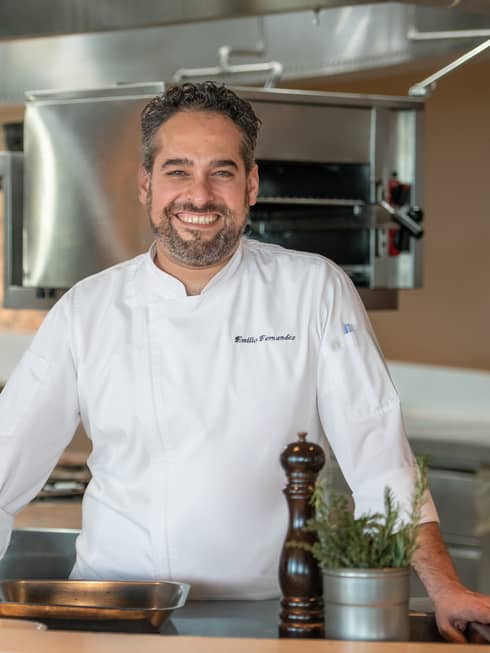 Executive Chef Emilio Fernandez in white chef's coat, standing in restaurant kitchen, smiling