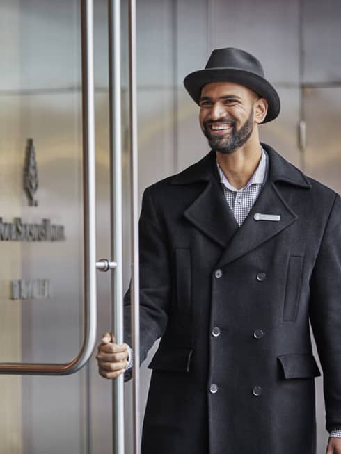 Smiling doorman in black coat, hat holds open front hotel lobby door
