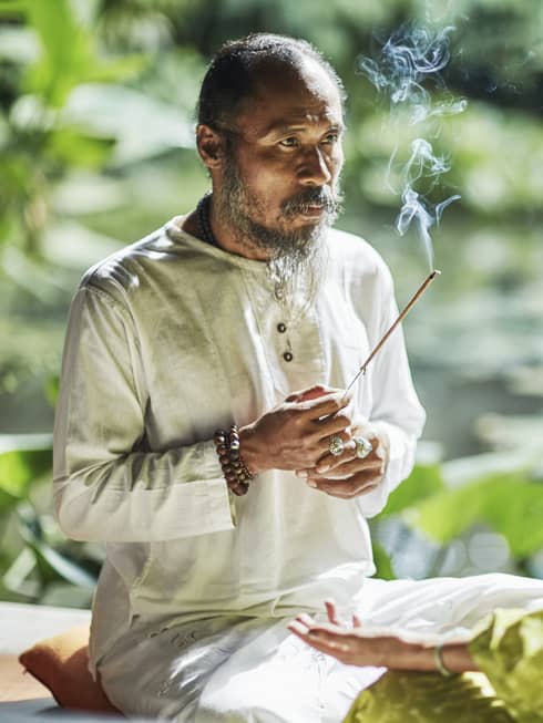 Set against a backdrop of greenery, Djik Dewa holds a stick of burning incense before a meditative cross-legged guest. 