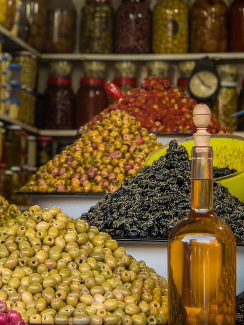 Bowls of colourful olives next to a smiling vendor holding out a scoop. Jars of preserves fill shelves in the background.