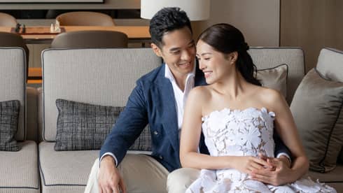 Smiling couple seated on a light gray sofa in a modern living room with decorative shelves and a large ocean artwork in the background.
