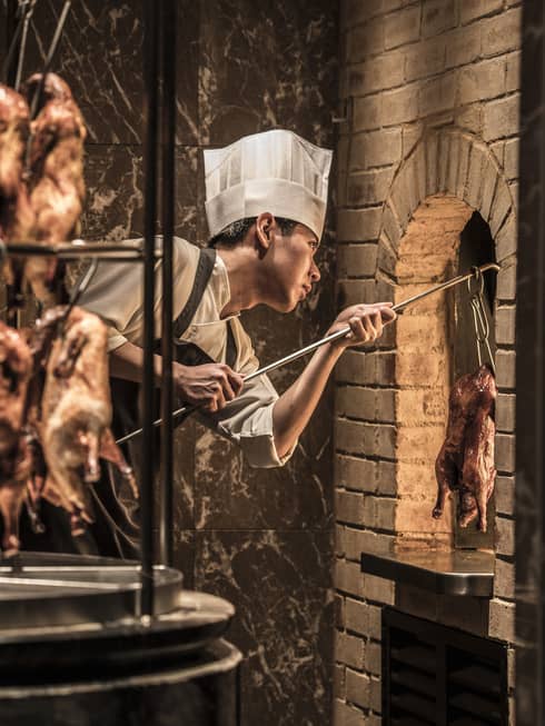 Rotisserie with hanging whole ducks in front of chef wearing white hat, taking duck from brick oven