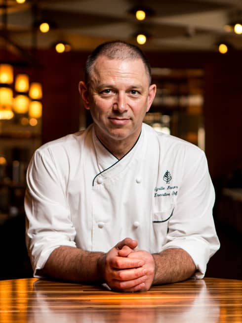 Portrait photo of Executive Chef Cyrille Pannier wearing white uniform, sitting at wood table with hands clasped in front